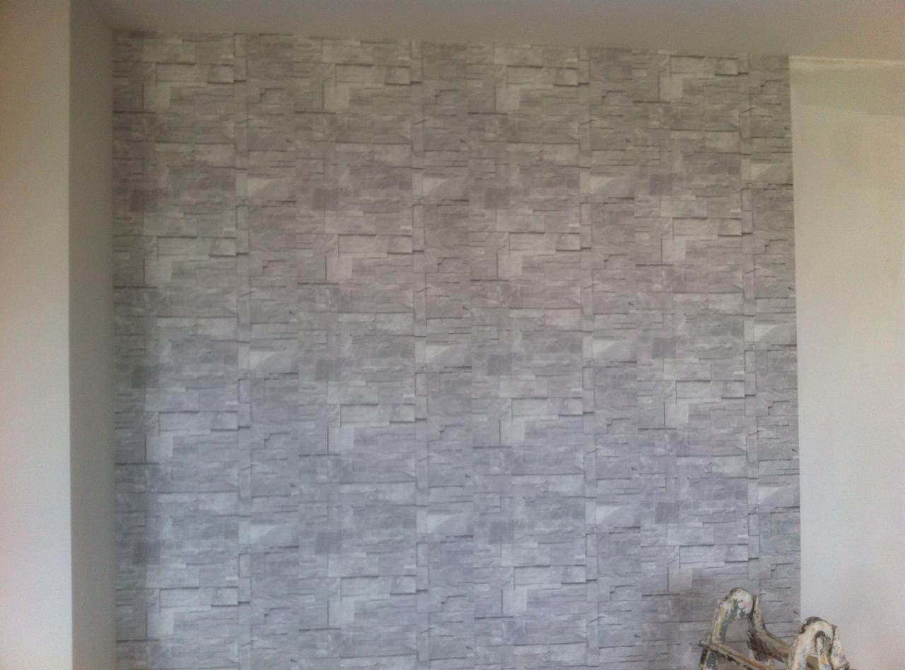 Ściana pokryta szarą tapetą imitującą kamień, widoczne łączenia pasów tapety, fragment drabiny malarskiej w rogu.