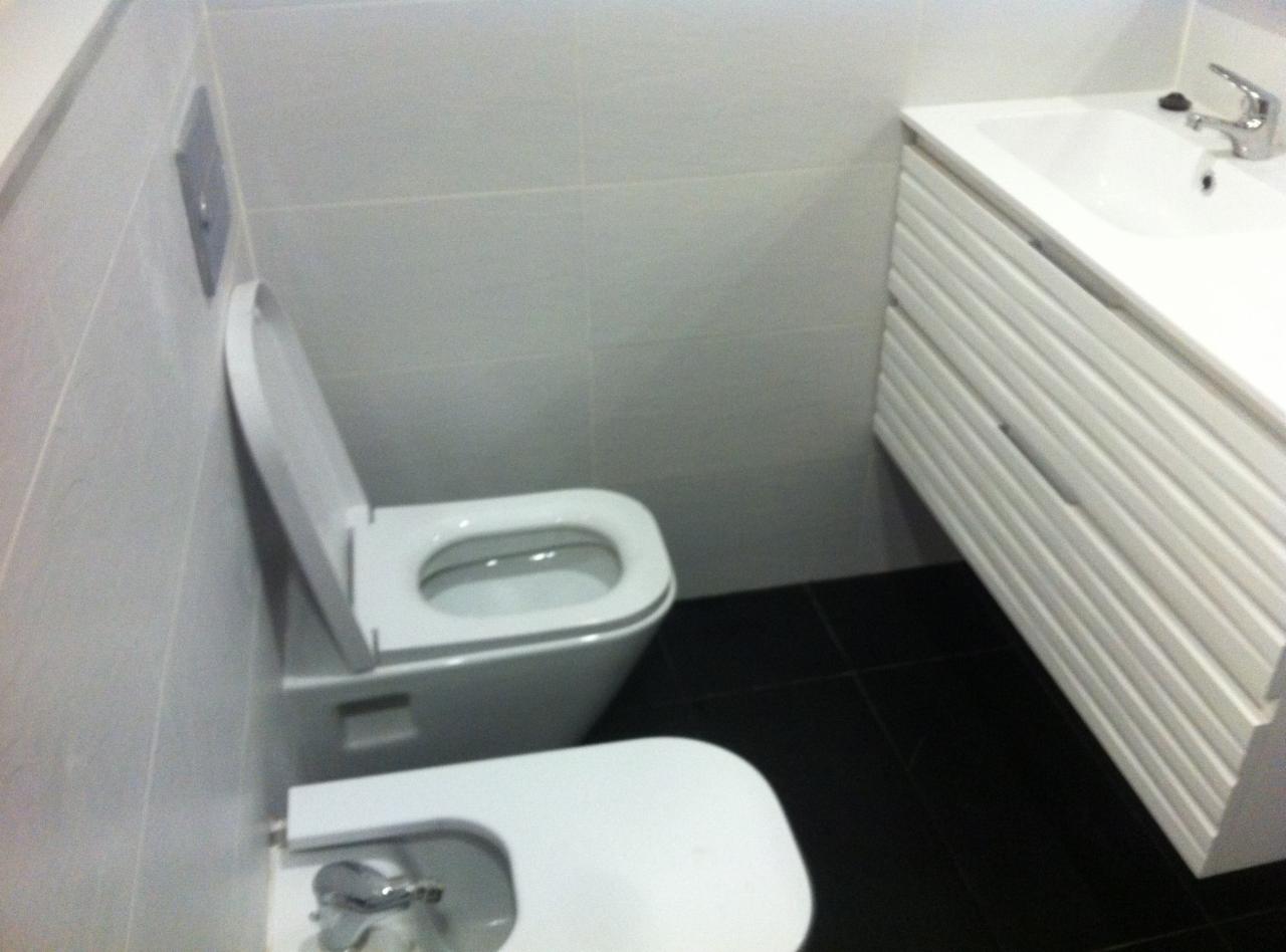 Nowoczesna łazienka z białą ceramiką: toaleta z otwartą klapą, bidet z chromowaną armaturą i biała szafka pod umywalkę z ryflowanymi frontami na tle czarnych płytek podłogowych i jasnych płytek...