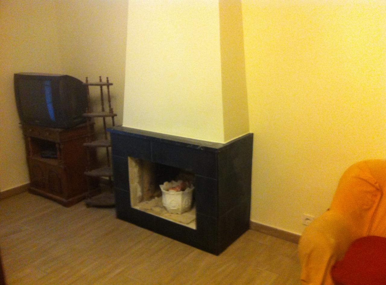 Narożny kominek obudowany czarnymi płytkami, wnętrze wyłożone beżowym kamieniem, ozdobione białym koszykiem z kwiatami, obok drewniany regał i telewizor kineskopowy