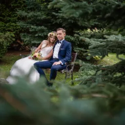 Uśmiechnięta para młoda w strojach ślubnych siedzi na metalowej ławce w otoczeniu zielonych drzew iglastych, panna młoda ma wianek na głowie i trzyma bukiet.