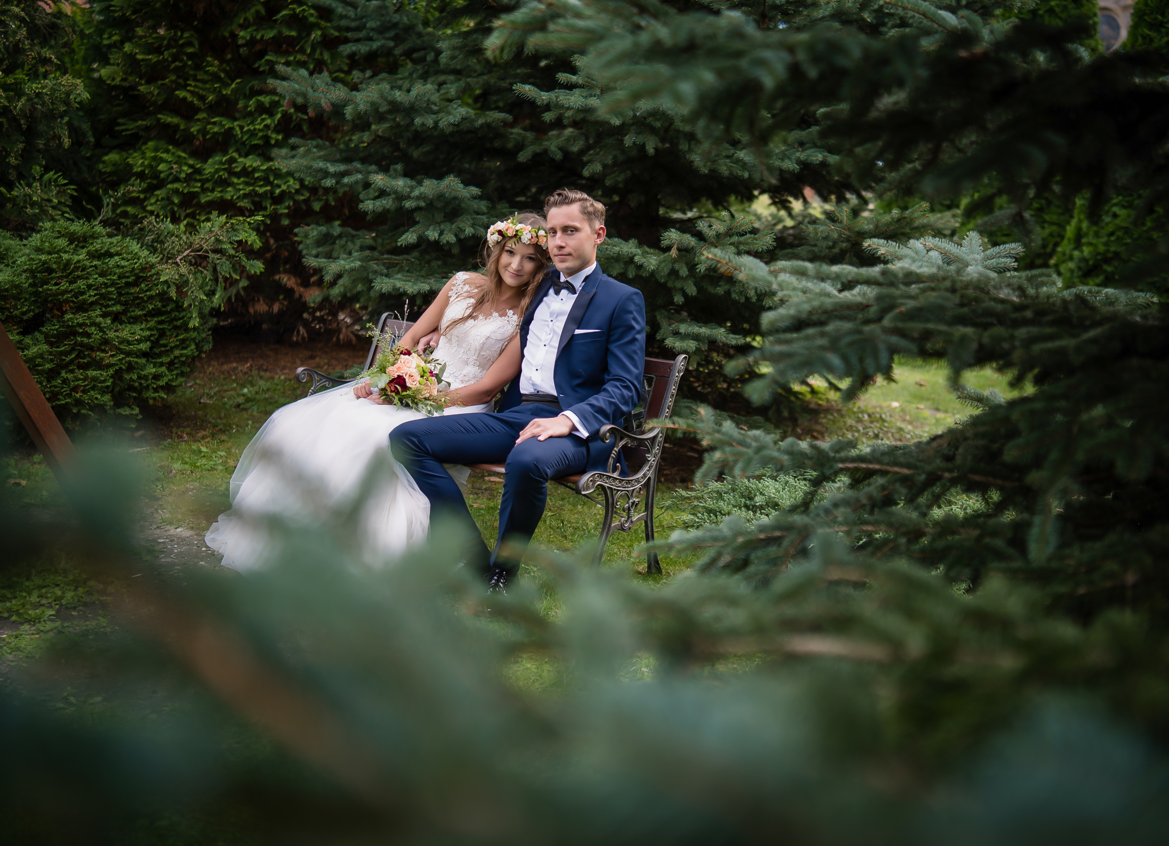 Uśmiechnięta para młoda w strojach ślubnych siedzi na metalowej ławce w otoczeniu zielonych drzew iglastych, panna młoda ma wianek na głowie i trzyma bukiet.