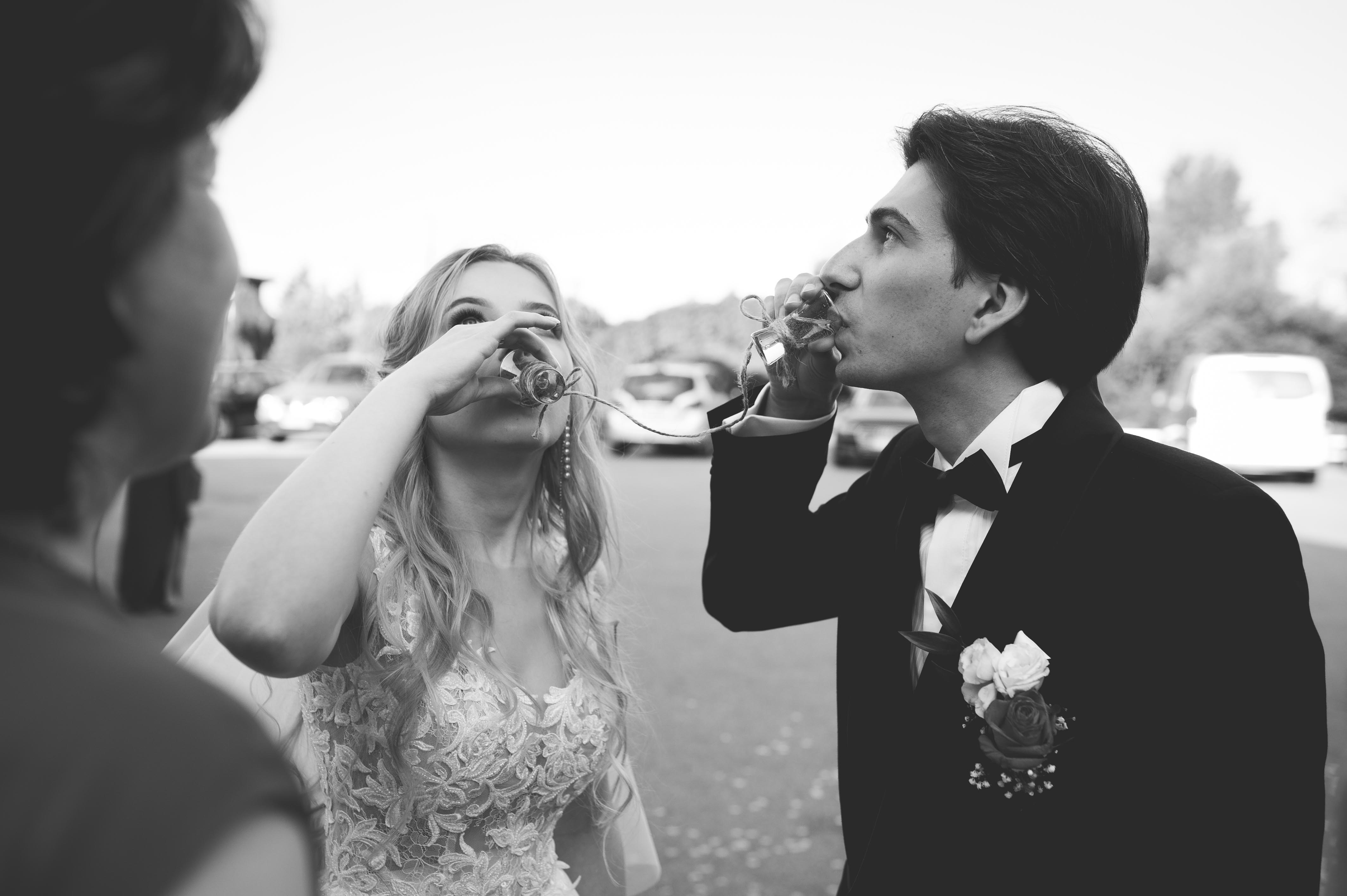 Czarno-białe ujęcie pary młodej wznoszącej toast małymi, kwadratowymi kieliszkami przewiązanymi sznurkiem, z widocznym bukietem butonierki pana młodego i koronkową suknią panny młodej.