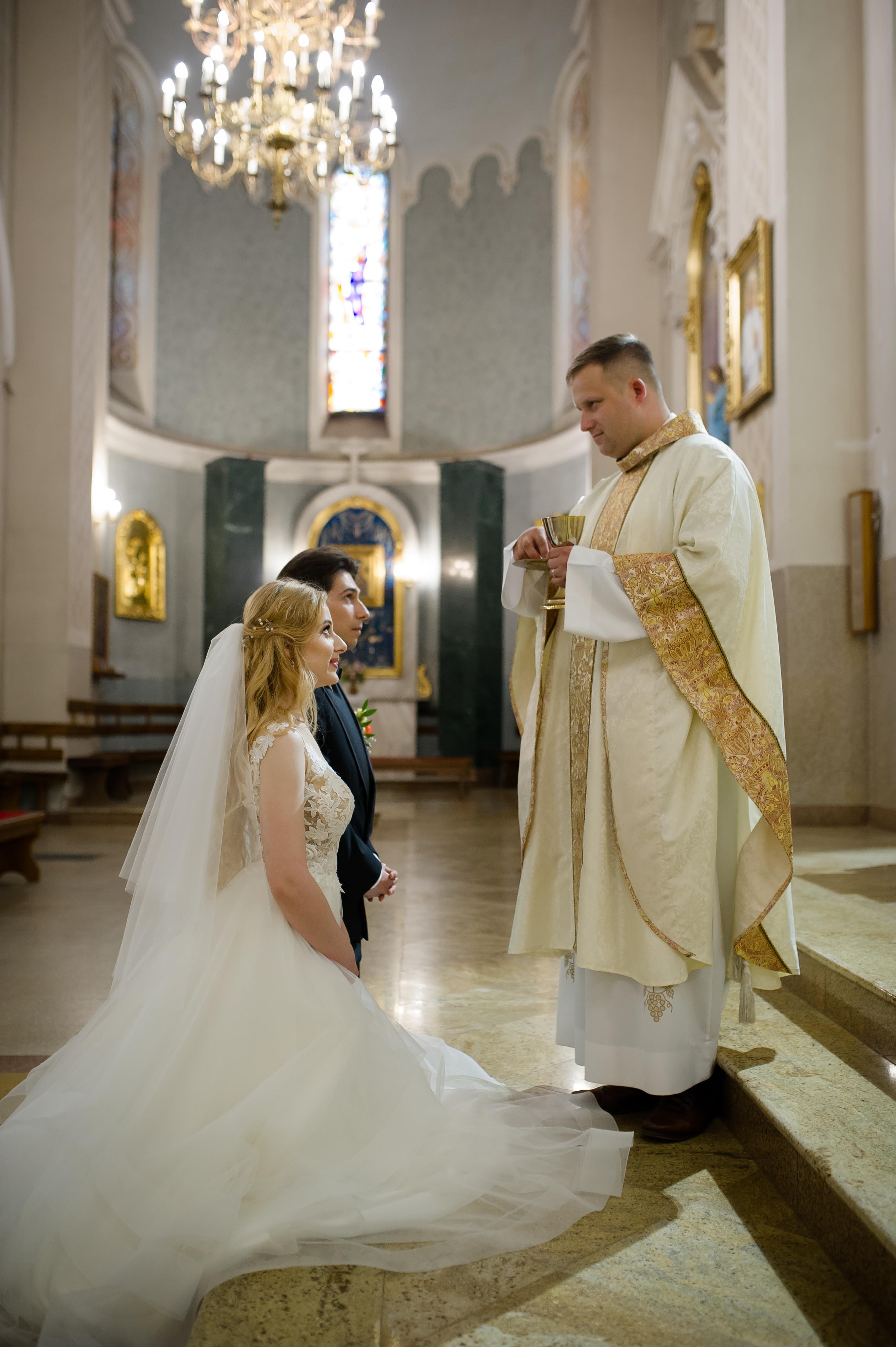Para młoda podczas ceremonii ślubnej w kościele, ksiądz błogosławi kielichem, w tle witraże i żyrandol.
