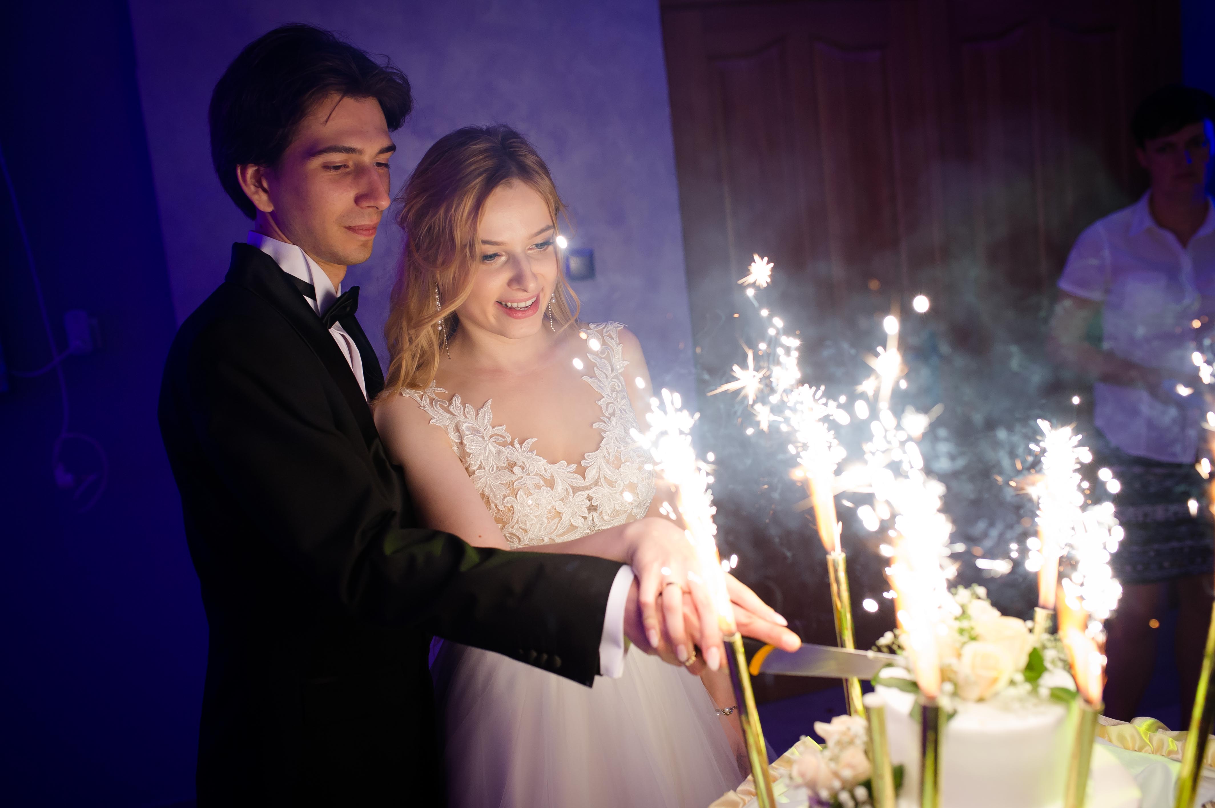 Młoda para w trakcie krojenia tortu weselnego z fontannami iskier, on w smokingu, ona w białej sukni z koronkową górą, w tle osoba obsługi.