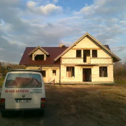 Niedokończony dom z czerwonym dachem, widoczny zza białego busa z numerem telefonu i napisem 'kompleksowe wykonawstwo pokryć dachowych'.