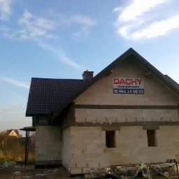 Niedokończony dom w Żyrzynie z brązowym dachem i widocznym banerem reklamowym firmy dekarskiej na szczycie ściany. Na pierwszym planie materiały budowlane.