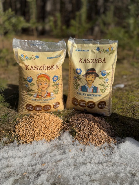 Dwa worki pelletu drzewnego Kaszëba z wizerunkami postaci w strojach kaszubskich, na tle leśnym i resztek śniegu. Jeden worek z dziewczyną, drugi z chłopakiem.