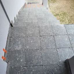 Schody zewnętrzne wyłożone szarą, kamienną płytką, widoczne pomarańczowe kliny dystansowe i krzyżyki do fug, perspektywa z góry.