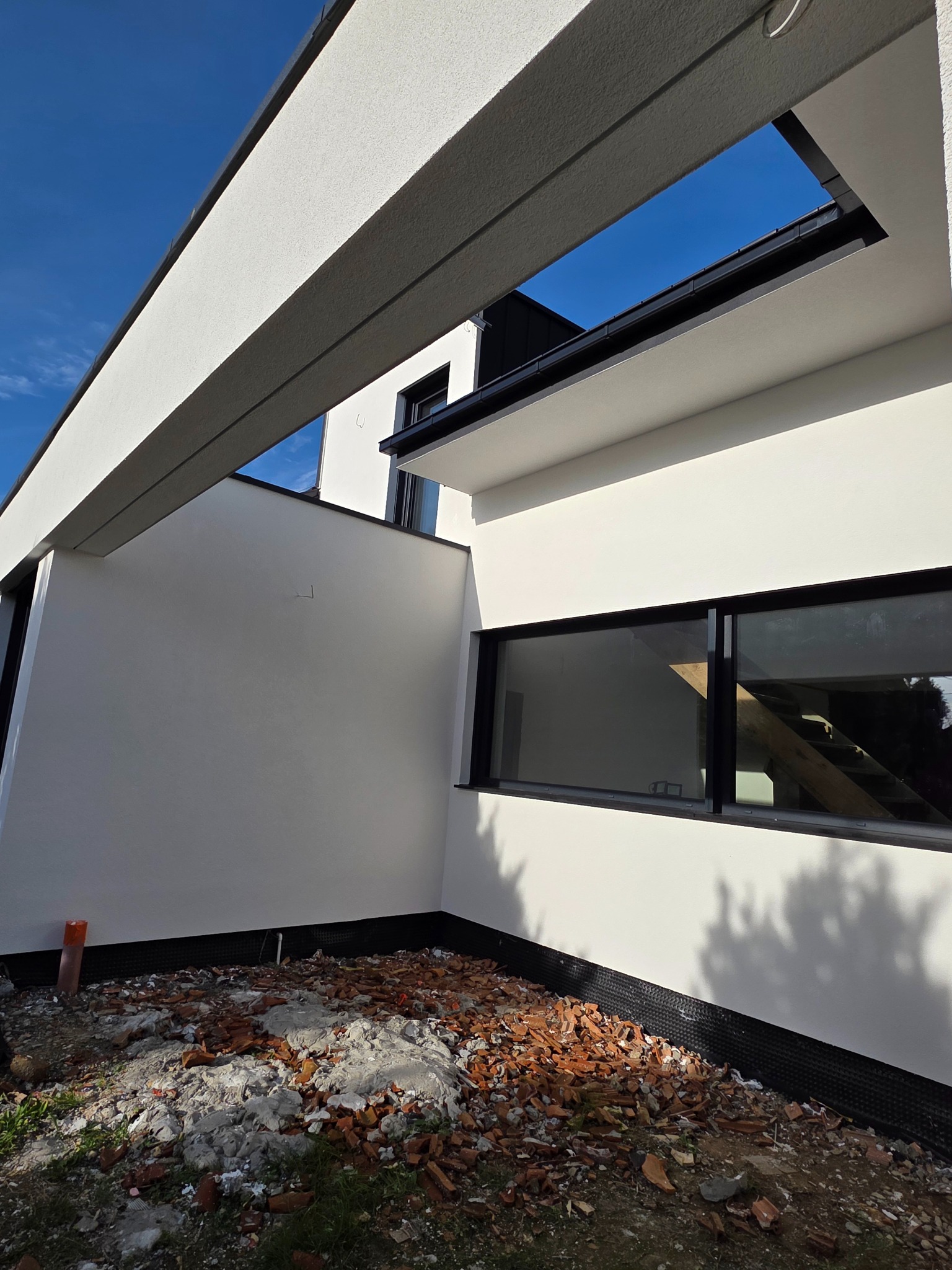 Biała elewacja domu w nowoczesnym stylu, widok z dołu na fragment ściany z oknem i elementami konstrukcyjnymi. Na ziemi gruz i resztki materiałów budowlanych.