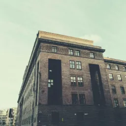 Wysoki, ceglany budynek z dużymi oknami i charakterystycznymi, pionowymi wnękami, widziany z dołu z perspektywy ulicy z przejściem dla pieszych.