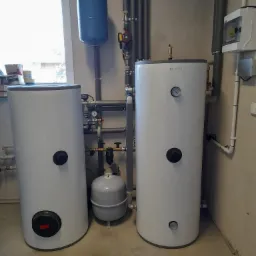 Dwa białe zasobniki ciepłej wody użytkowej marki Roensol z podłączeniami hydraulicznymi, widoczne także naczynia wzbiorcze, rury i zawory w surowym pomieszczeniu.