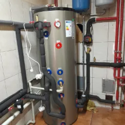 Szary bojler z licznymi rurami, zaworami i manometrem w kotłowni z beżowymi płytkami na ścianach i brązową terakotą na podłodze. Widoczne r&oacute;wnież dwa zbiorniki wyr&oacute;wnawcze nad bojlerem.