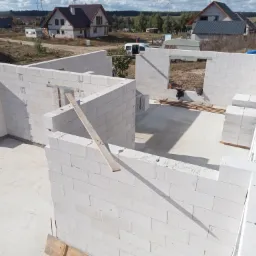 Budowa domu w trakcie: białe ściany z bloczków, widoczne wiązania, częściowo ułożone, w tle domy jednorodzinne i wiatraki.