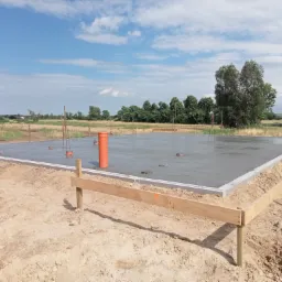 Wylany fundament płyty na gruncie, widoczna szalunkowa rama z drewna i pomarańczowa rura wentylacyjna, w tle łąka i drzewa pod błękitnym niebem.