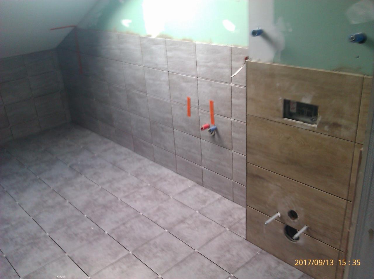 Łazienka w trakcie układania płytek: szare płytki ścienne i podłogowe, widoczne przyłącza wodne, fragment ściany z obudową stelaża podtynkowego WC.