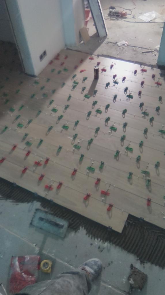 Układanie płytek podłogowych w pomieszczeniu, widoczne klipsy poziomujące, puszka po kleju, narzędzia i stopa w roboczym obuwiu.