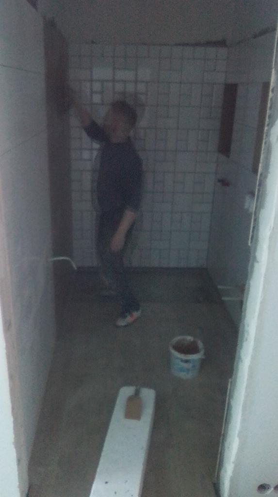 Pracownik w trakcie fugowania płytek w łazience, widoczny kubeł z fugą i poziomica na podłodze, słabe oświetlenie.