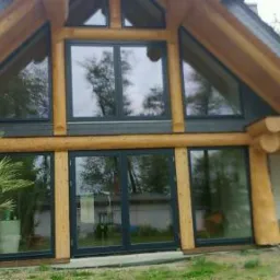 Dom z bali w stylu skandynawskim z dużymi przeszkleniami w ciemnych ramach, widoczne odbicia drzew w szybach, na pierwszym planie palma.