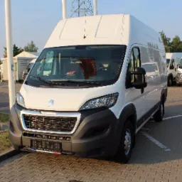 Biały bus Peugeot Boxer z wysokim dachem, zaparkowany na brukowanym placu, widok z przodu, w tle inne busy i budynki.