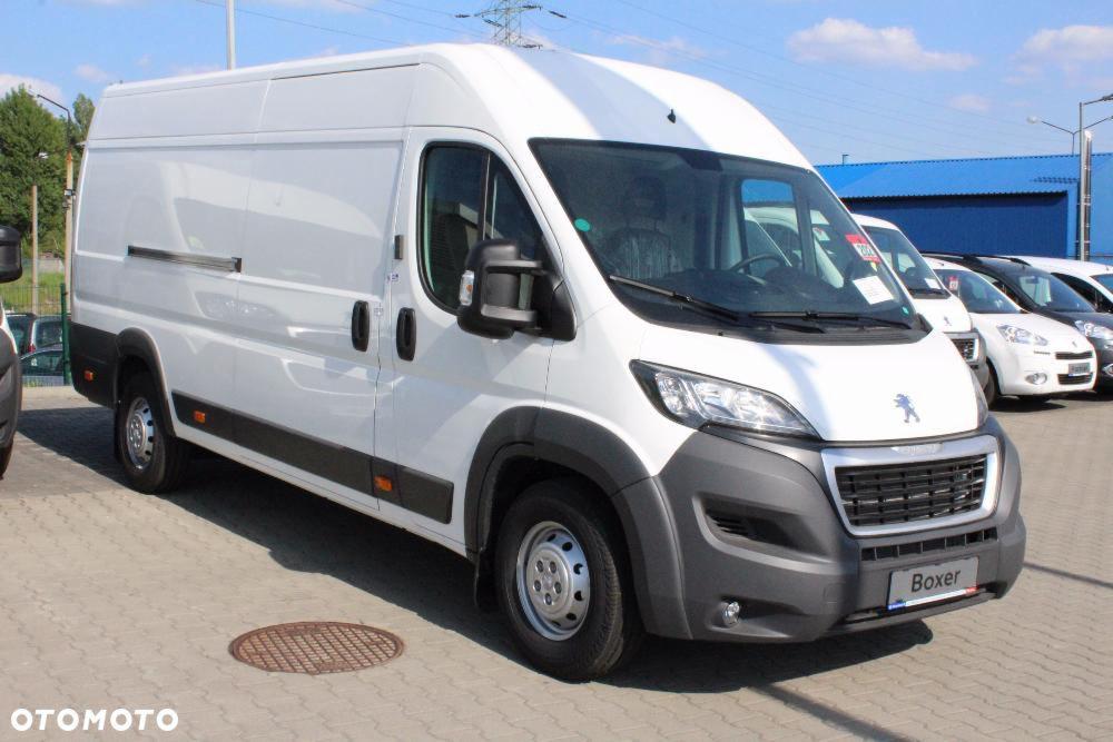Biały samochód dostawczy Peugeot Boxer na parkingu, widoczny z boku, z innymi samochodami w tle i studzienką kanalizacyjną na pierwszym planie.