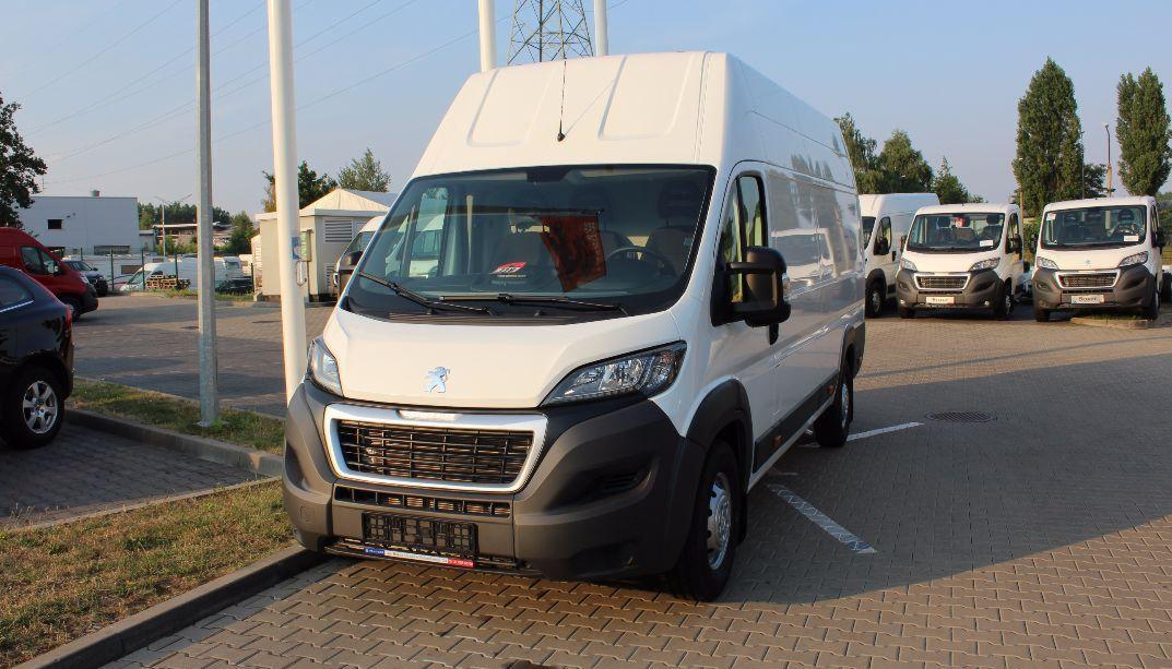 Biały bus Peugeot Boxer z wysokim dachem, zaparkowany na brukowanym placu, widok z przodu, w tle inne busy i budynki.
