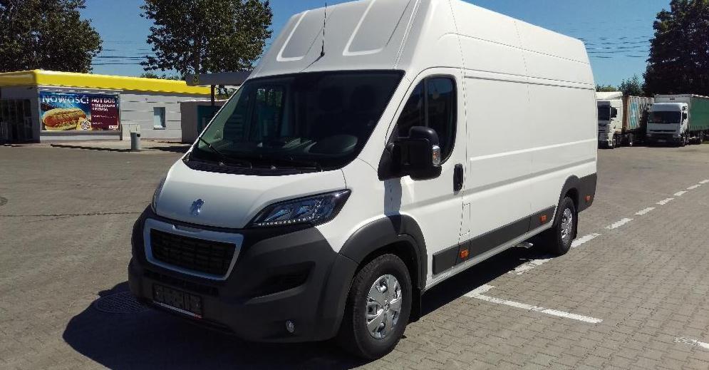 Biały bus dostawczy marki Peugeot zaparkowany na parkingu z widocznym budynkiem stacji benzynowej w tle.