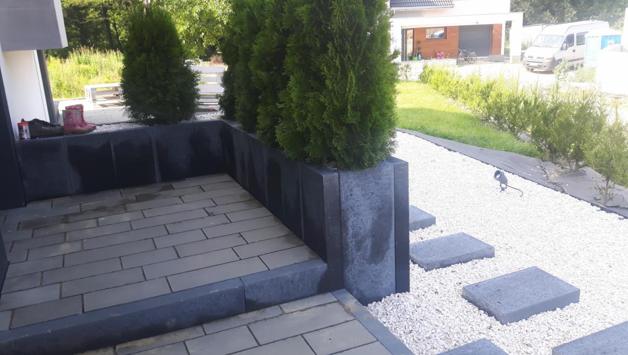 Ułożona szara kostka brukowa w kształcie cegieł tworząca taras z donicami z tujami, obok ścieżka z białego żwiru z kamiennymi płytami, w tle dom jednorodzinny i zaparkowany van.