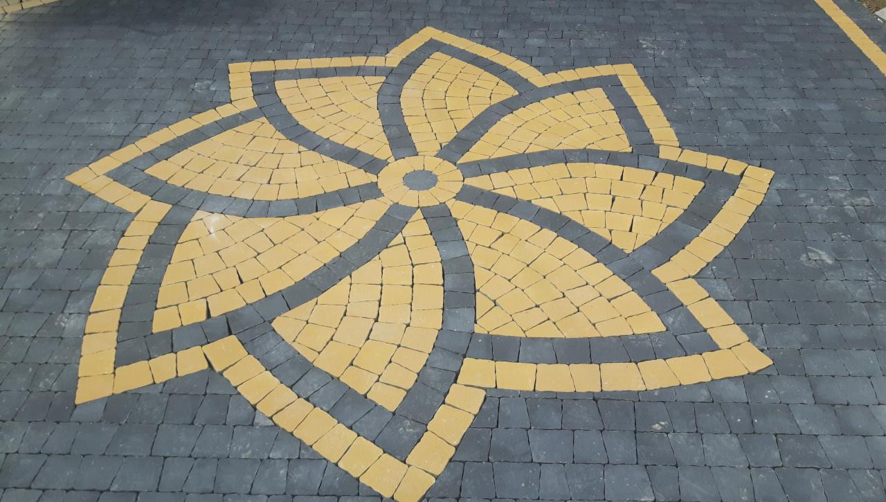 Wzór kwiatu ułożony z żółtej kostki brukowej na tle szarej kostki, widok z góry.