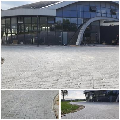Szara nawierzchnia z kostki ułożona w łukowaty wzór przed nowoczesnym budynkiem z przeszkloną fasadą. Widoczne detale krawężnika i trawnika w oddali.