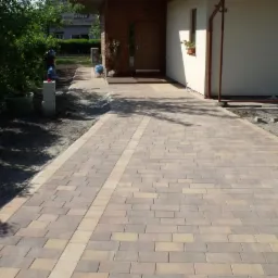 Ułożona nawierzchnia z kostki brukowej w odcieniach beżu i brązu prowadząca do wejścia domu, widoczne przygotowane obrzeża i fragmenty niezabudowanego terenu.