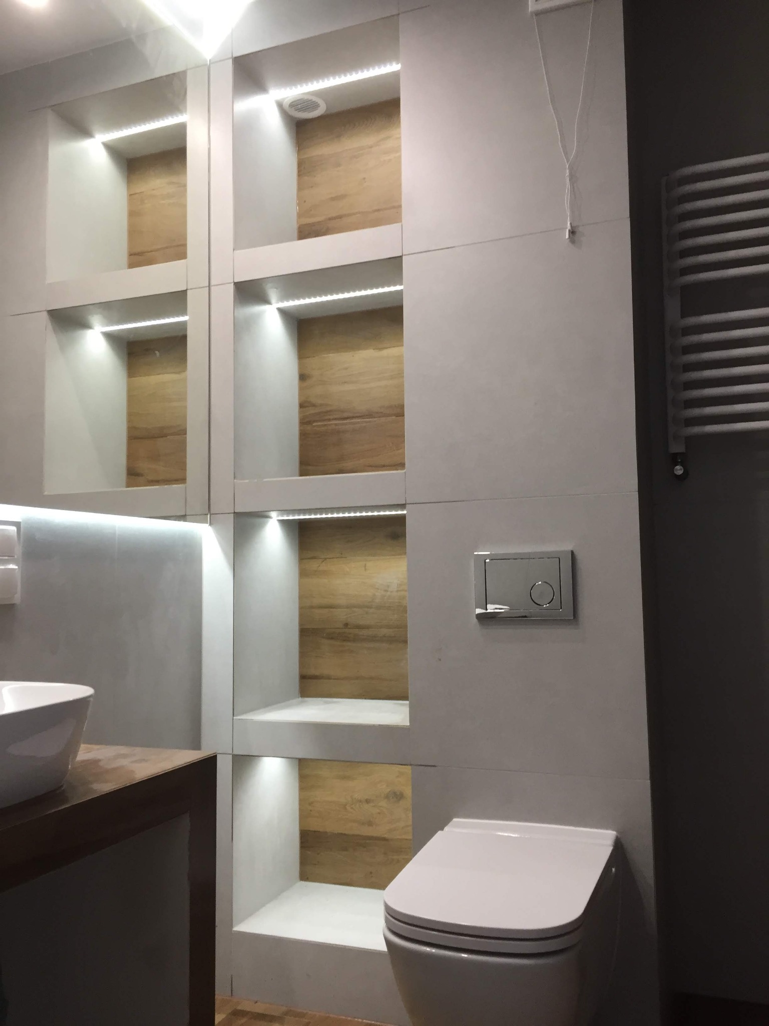 Nowoczesna łazienka z podświetlanymi wnękami wyłożonymi drewnem, umywalką nablatową i wiszącą toaletą.