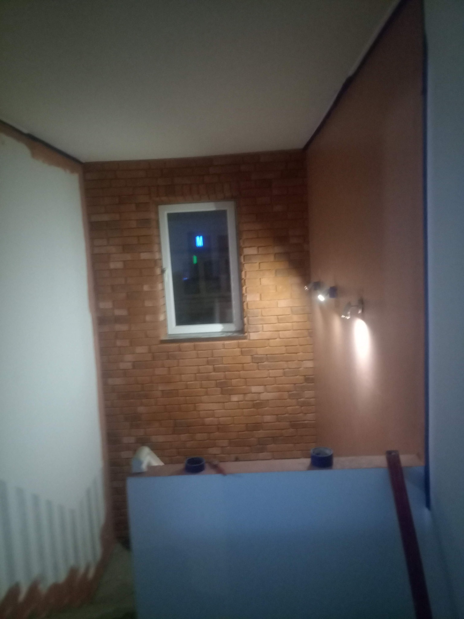 Wnętrze w trakcie wykańczania: ściana z cegły klinkierowej z oknem, gładka ściana z oświetleniem punktowym, widoczne narzędzia i materiały budowlane, brak wykończenia podłogi i sufitu.