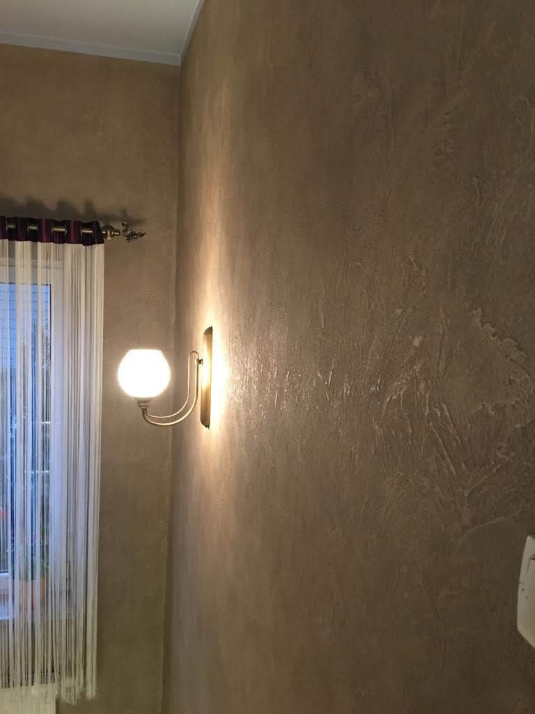 Ściana z dekoracyjnym tynkiem w odcieniach brązu i beżu, oświetlona przez kinkiet z okrągłą, mleczną żarówką, obok okna z firanką.