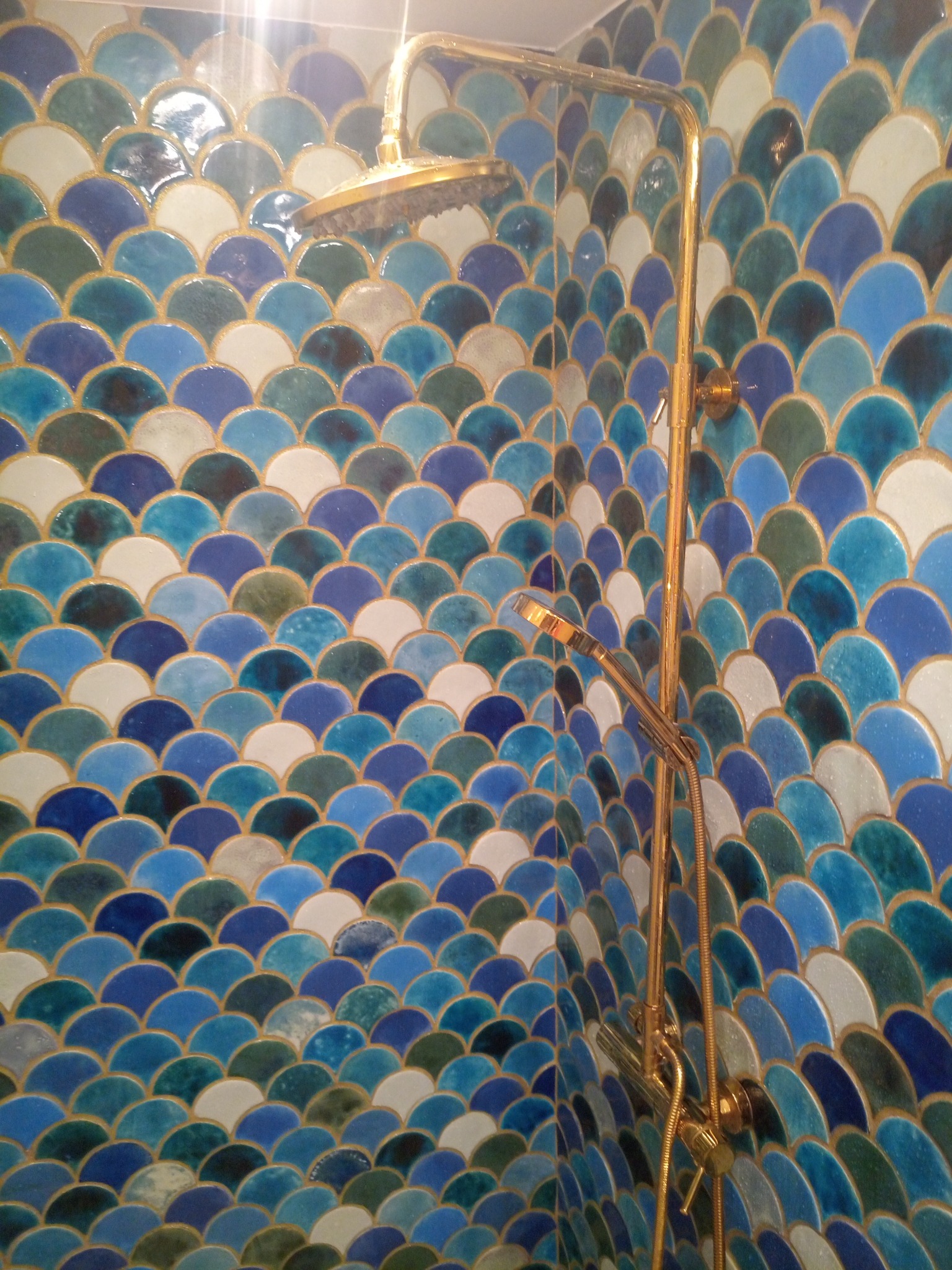 Ściana prysznica wyłożona mozaiką w kształcie rybiej łuski w odcieniach błękitu, turkusu i bieli, z złotą armaturą prysznicową.