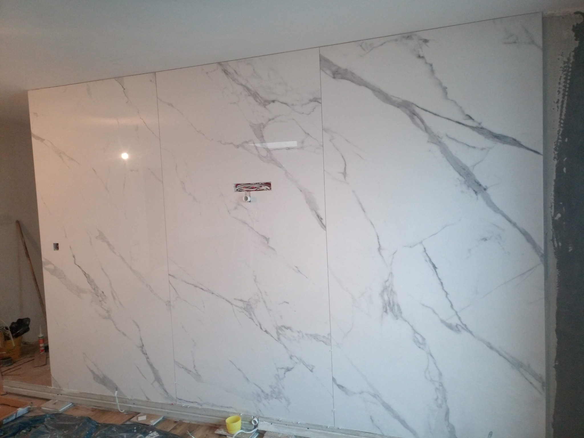 Ściana wyłożona wielkoformatowymi płytami imitującymi marmur, widoczne przewody elektryczne przygotowane do montażu gniazdka, fragmenty narzędzi i materiałów budowlanych na podłodze.