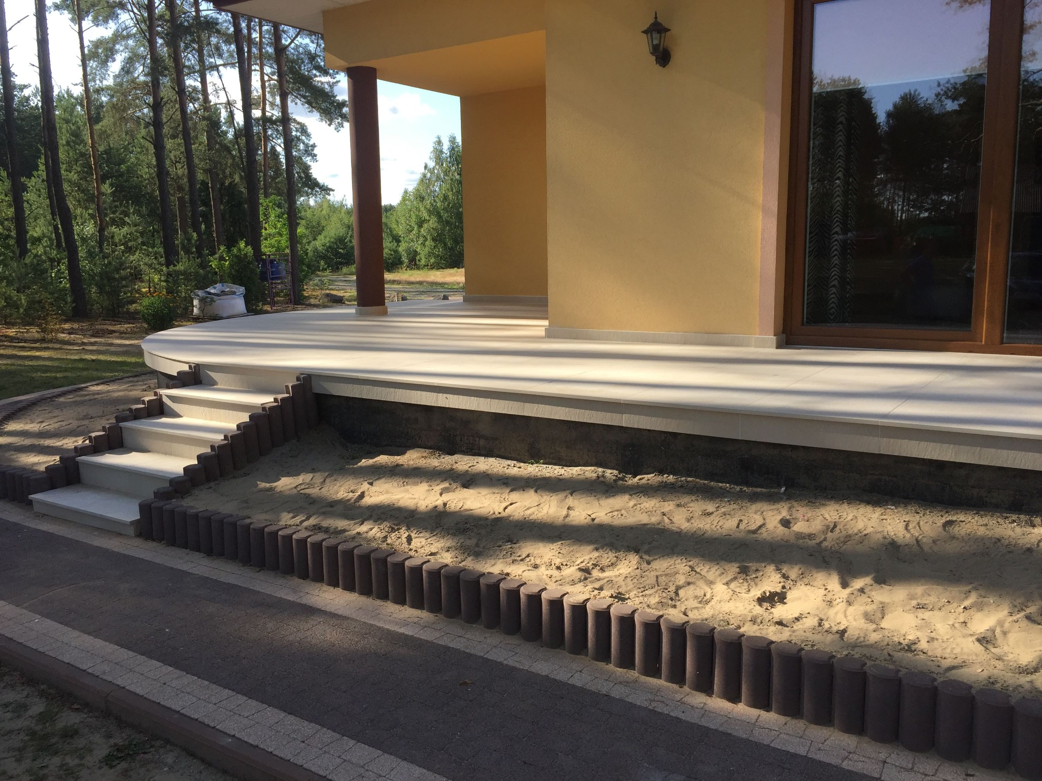 Wykończony taras z jasnymi płytkami, schody z jasnymi płytkami i obrzeże z brązowych palisad, z widokiem na las w tle.