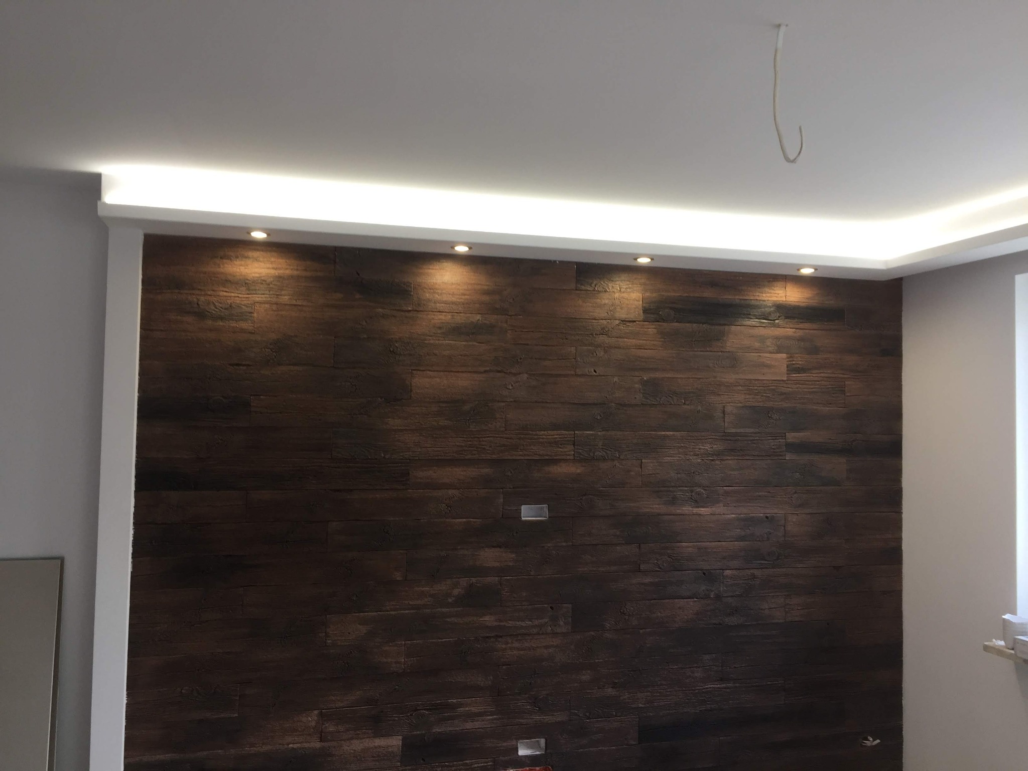 Ściana wykończona panelami imitującymi ciemne drewno, z wbudowanym oświetleniem punktowym i listwą LED na suficie, widoczne niedokończone podłączenie elektryczne na suficie.