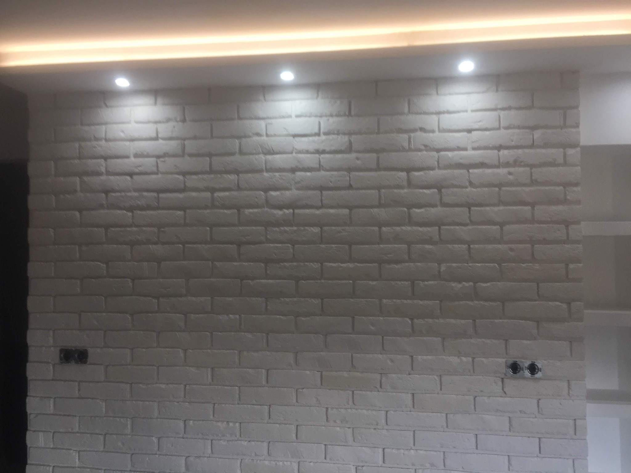 Biała ściana z cegły dekoracyjnej podświetlona od góry trzema punktowymi lampami, z widocznymi gniazdkami elektrycznymi.