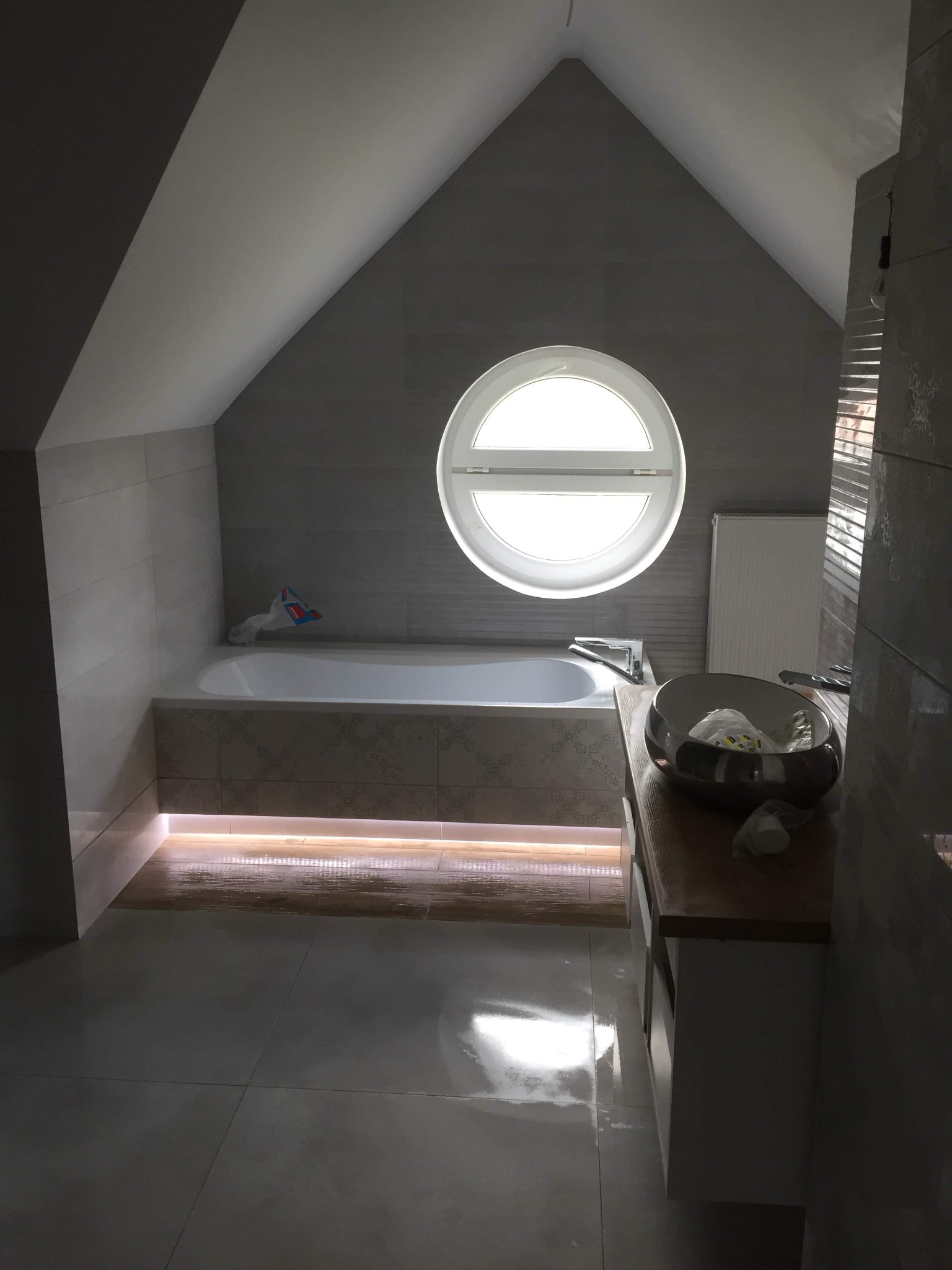 Nowoczesna łazienka na poddaszu z okrągłym oknem, szarymi płytkami, podświetlaną wanną, umywalką na drewnianym blacie i błyszczącą podłogą.