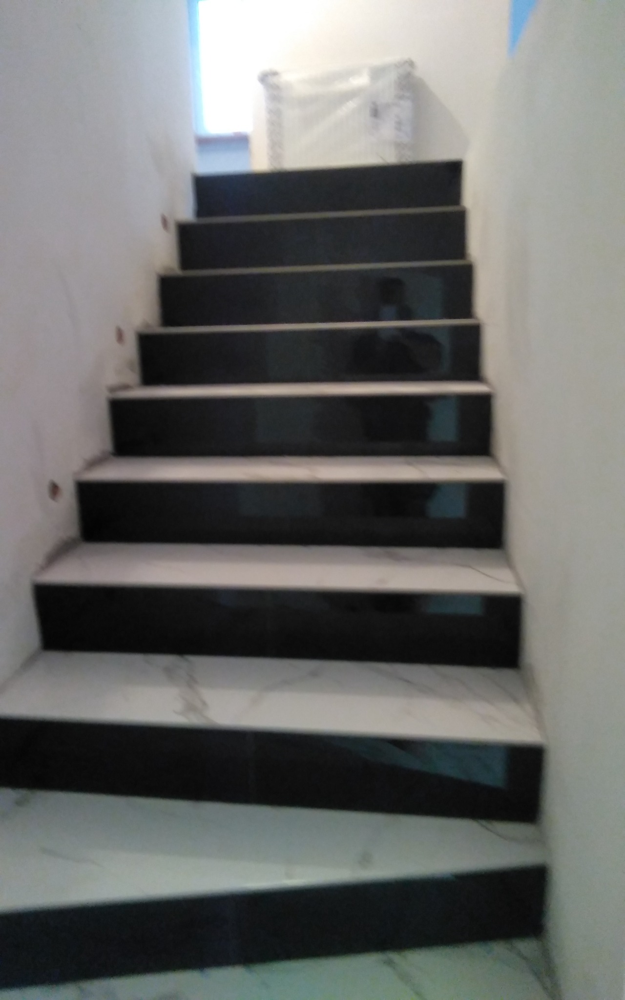 Schody wyłożone czarno-białymi płytkami, widoczne z dołu, z białymi ścianami po bokach i oknem w tle.