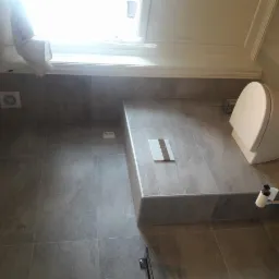 Wykończona łazienka z szarymi płytkami, podwieszaną toaletą i oknem z roletą. Widoczny grzejnik wentylatorowy na ścianie.