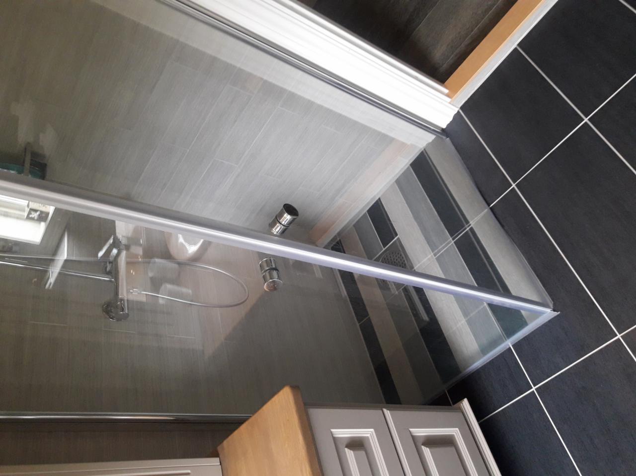 Nowoczesna kabina prysznicowa z szklanymi drzwiami przesuwnymi i chromowanymi uchwytami, szare płytki imitujące drewno na ścianach prysznica, kontrastujące czarne płytki podłogowe z białymi fugami,...