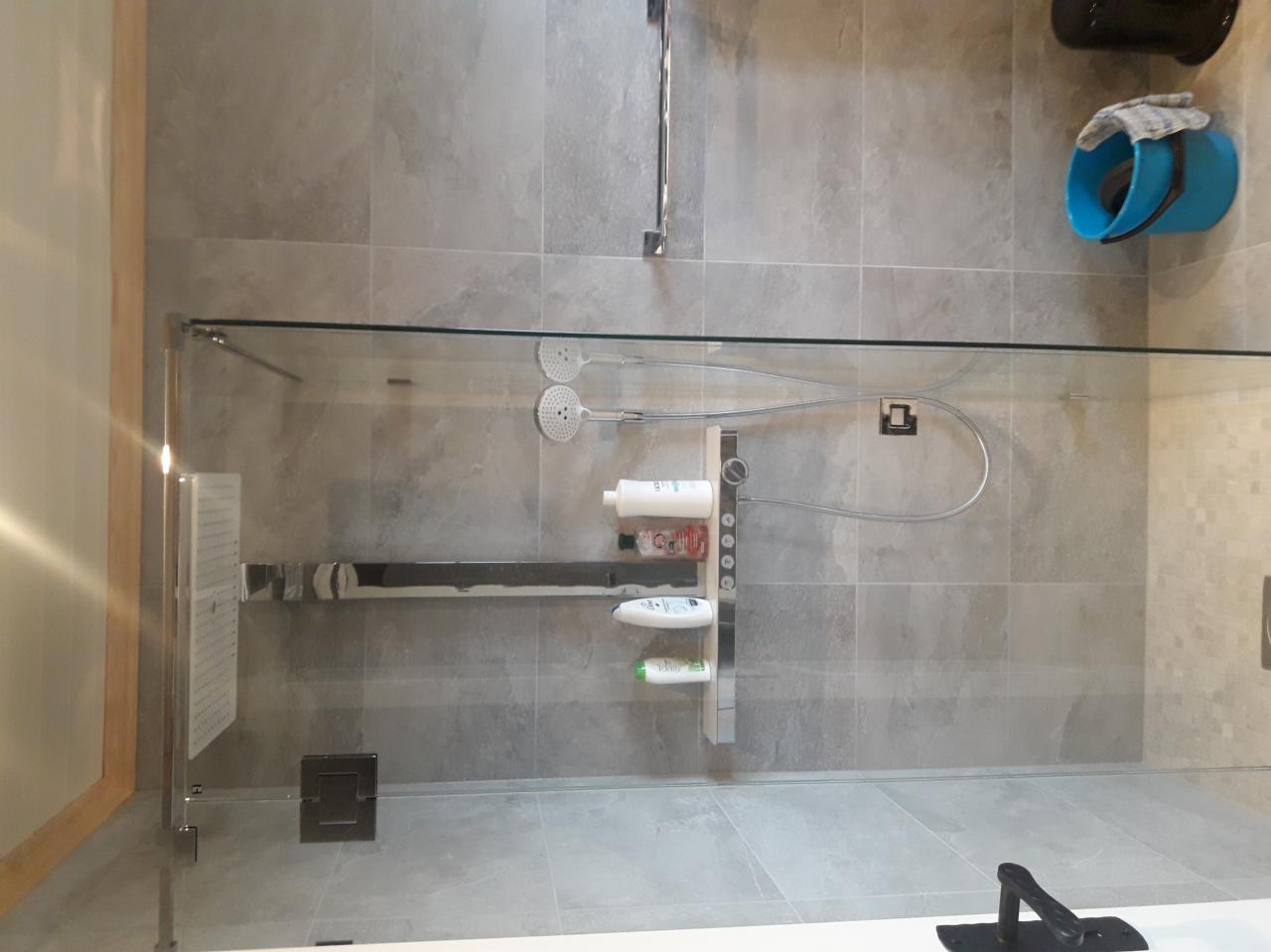 Szklana kabina prysznicowa z szarymi płytkami, półką na kosmetyki i dwoma słuchawkami prysznicowymi.