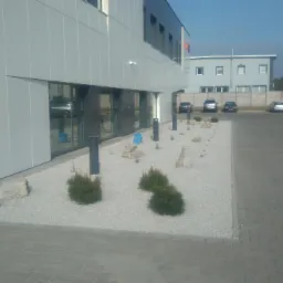 Nowoczesny budynek biurowy z minimalistycznym ogrodem: jasne kamienie, niskie krzewy iglaste, pojedyncze kamienie dekoracyjne, oświetlenie punktowe wzdłuż elewacji, zaparkowane samochody w tle.