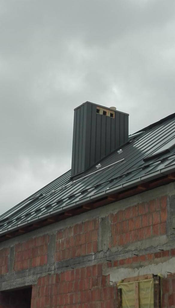 Szary blaszany dach z kominem w trakcie budowy, widoczny surowy mur z czerwonej cegły i pochmurne niebo.