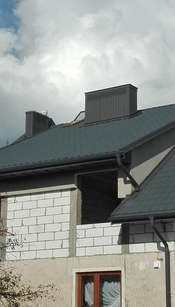 Nowy, grafitowy dach z blachodachówki na surowym budynku w trakcie budowy, widoczne kominy wentylacyjne i niezabudowany otwór okienny.