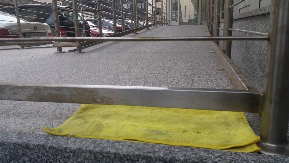 Żółta ściereczka leżąca na szarym, granitowym cokole rampy dla niepełnosprawnych, z metalową barierką. Widoczny fragment czyszczonej powierzchni i zaparkowane samochody w tle.