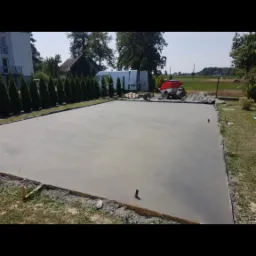 Świeżo wylana, gładka posadzka na zewnątrz, z widocznymi deskami szalunkowymi i sprzętem budowlanym w tle, otoczona zielenią.