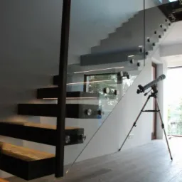 Nowoczesne schody z drewnianymi stopniami i szklaną balustradą, mocowaną za pomocą metalowych uchwyt&oacute;w; na podłodze widoczny teleskop.