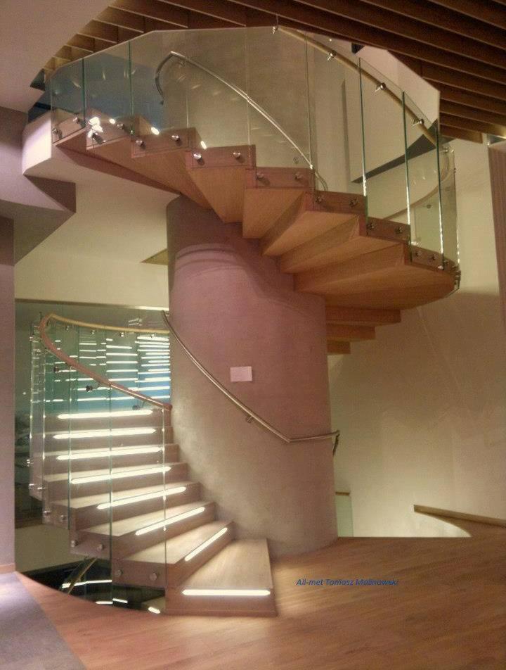 Spiralne schody z drewnianymi stopniami i szklaną balustradą, podświetlone LED-ami, umieszczone wokół betonowego filaru.